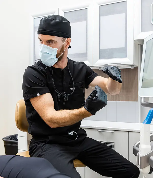 A dentist wearing a mask and gloves sits in an exam room explaining a dental procedure to a patient, using hand gestures to illustrate a concept.