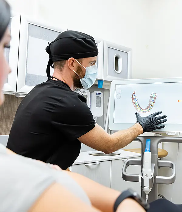 Dentist wearing scrubs, gloves, and a mask explains a digital dental scan to a patient using a monitor in a modern dental office.