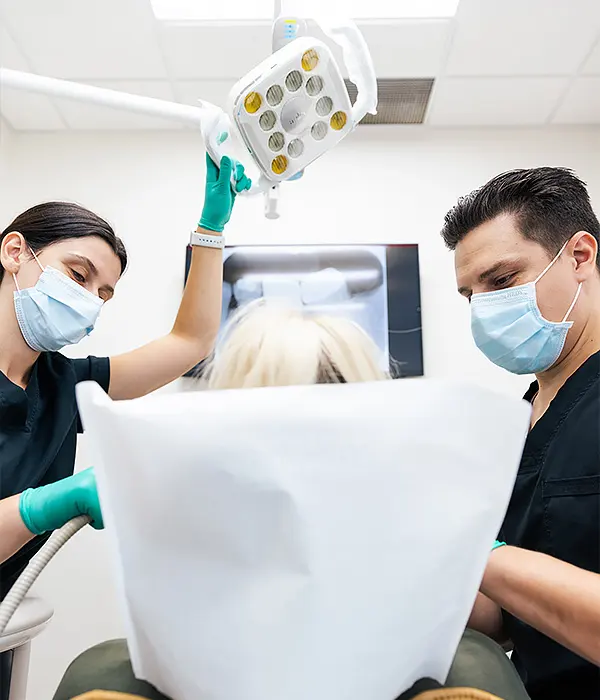 Two masked dental professionals prepare to examine or treat a patient lying in a dental chair, adjusting an overhead light in a clean, modern clinic.