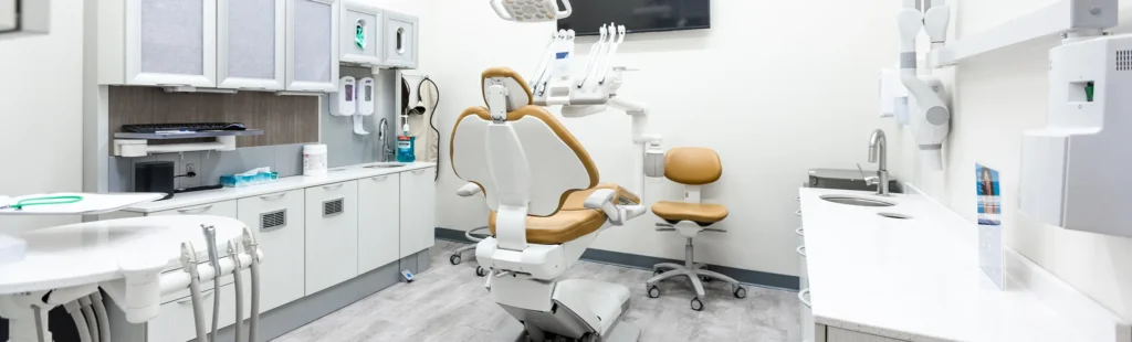 Modern dental treatment room with clean white cabinetry, tan dental chairs, mounted equipment, and sterilization supplies, creating a bright and organized clinical space.
