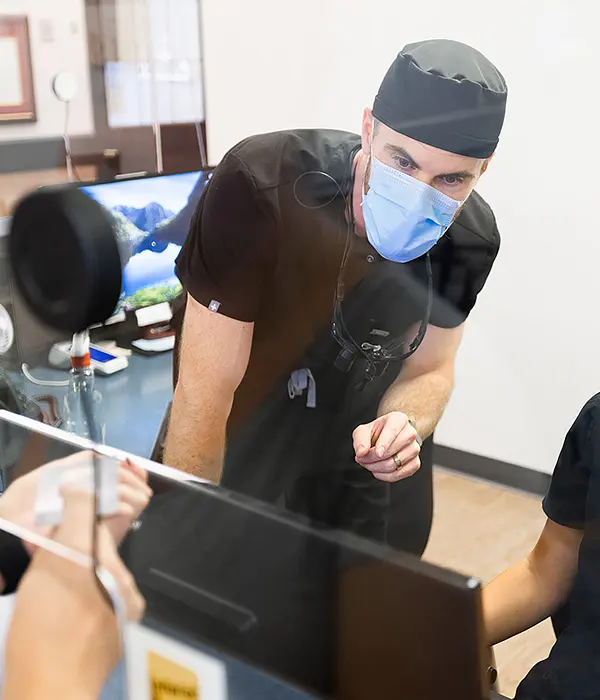 Dentist wearing a mask and scrubs leans over a reception desk, discussing appointment scheduling with a team member behind a transparent partition.